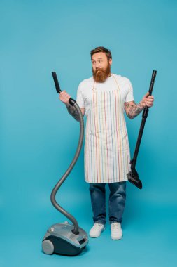 full length of discouraged man in striped apron looking at parts of vacuum cleaner on blue background