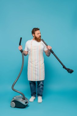 full length of thoughtful man in striped apron looking in tube of vacuum cleaner on blue background