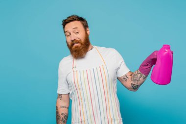 smiling tattooed man in striped apron holding bottle with washing gel isolated on blue