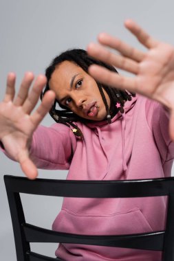 pierced multiracial man in pink hoodie gesturing while sitting on chair isolated on grey 