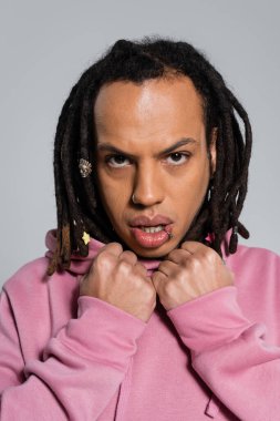 portrait of pierced multiracial man in pink hoodie looking at camera isolated on grey 