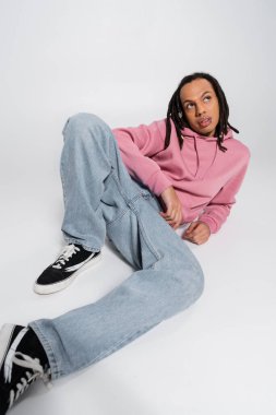full length of multiracial and pierced man with dreadlocks looking up while resting on grey 