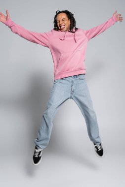full length of excited multiracial man with dreadlocks jumping with outstretched hands on grey 