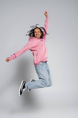 full length of positive multiracial man with dreadlocks jumping with outstretched hands on grey  clipart