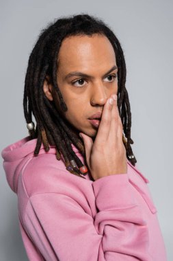 multiracial man with dreadlocks and piercing covering mouth with hand isolated on grey 