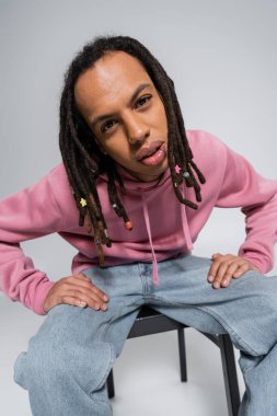 multiracial man with dreadlocks and piercing looking at camera while sitting on chair isolated on grey 