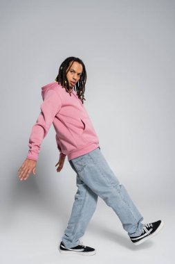 full length of multiracial man in blue jeans and pink hoodie posing on grey 