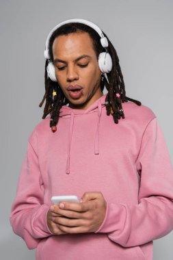 shocked multiracial man with dreadlocks using smartphone while listening music in wireless headphones isolated on grey 