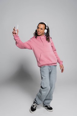 full length of multiracial man with dreadlocks taking selfie while listening music in wireless headphones on grey 