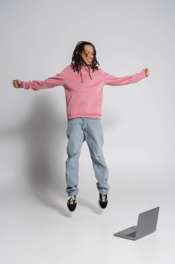 full length of happy multiracial man in pink hoodie and jeans jumping near laptop on grey 