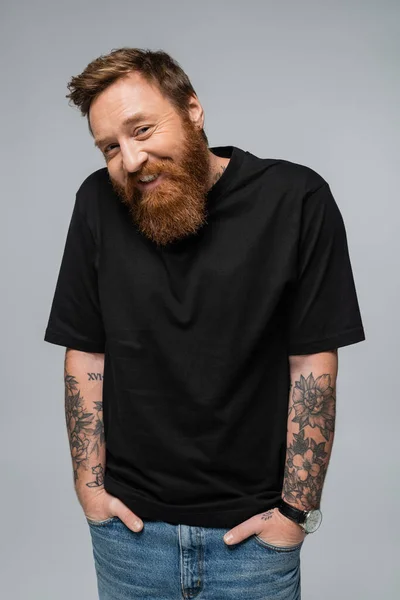 stock image cheerful bearded man in black t-shirt standing with hands in pockets of blue jeans isolated on grey