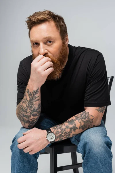 pensive tattooed man in black t-shirt and wristwatch holding hand near face and looking at camera isolated on grey