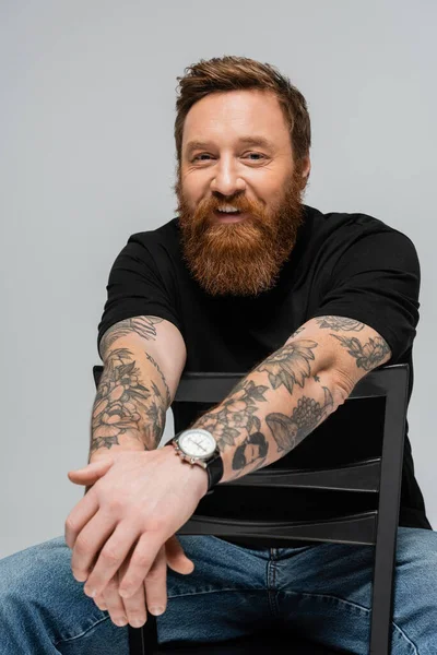 bearded tattooed man in black t-shirt and wristwatch sitting on chair and smiling at camera isolated on grey