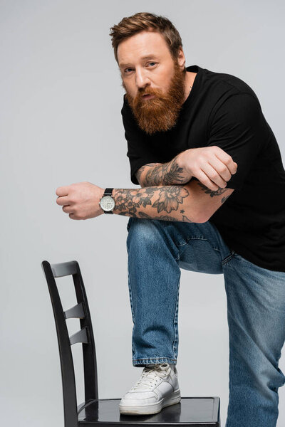 бородатый татуированный мужчина в синих джинсах и кроссовках, шагающий на стул и смотрящий на камеру, изолированную на сером