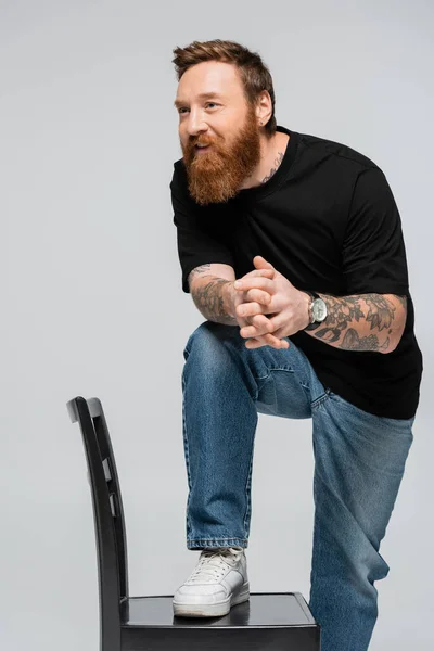 smiling man in black t-shirt and jeans looking away while stepping on chair isolated on grey
