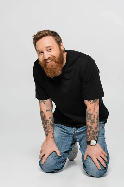 happy tattooed man in black t-shirt and jeans smiling at camera on grey background