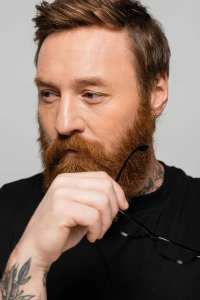 thoughtful bearded man holding eyeglasses and looking away isolated on grey