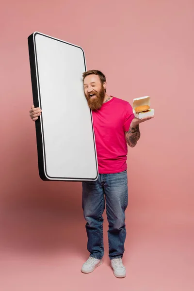 stock image bearded man holding hamburger and huge template of mobile phone while laughing with closed eyes on pink background