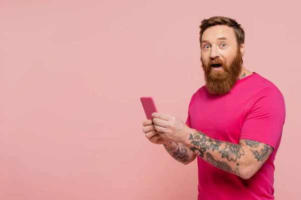 excited bearded man with open mouth using cellphone and looking at camera isolated on pink