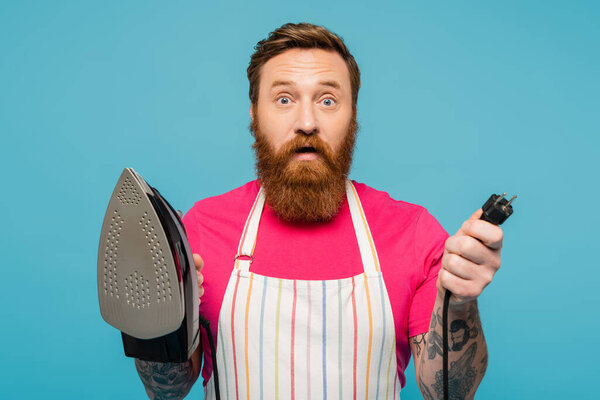discouraged bearded man in striped apron holding iron and looking at camera isolated on blue