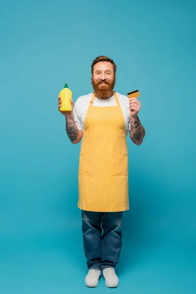 full length of happy bearded man in polka dot apron posing with credit card and detergent on blue background