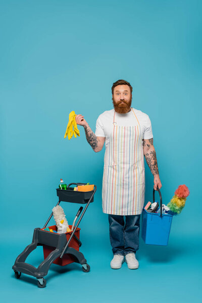 positive bearded man with rubber gloves and bucket looking at camera near cart with cleaning supplies on blue background