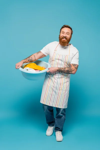 full length of happy tattooed man in striped apron holding laundry bowl and smiling at camera on blue background