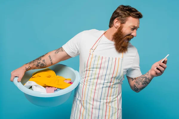 stock image shocked man in apron screaming while holding laundry bowl and looking at smartphone isolated on blue