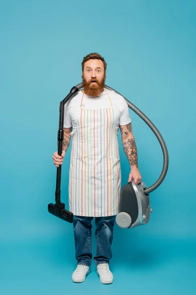 full length of discouraged man in apron holding vacuum cleaner and looking at camera on blue background