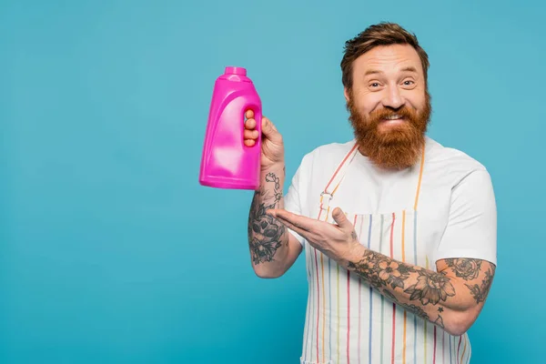 Stock image joyful bearded man in striped apron pointing at bottle with washing gel isolated on blue