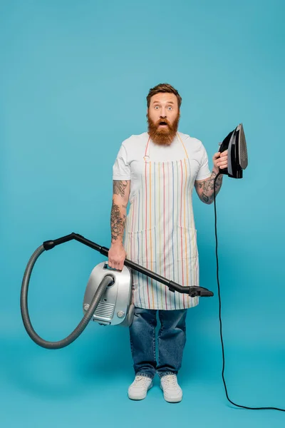 full length of shocked man in apron holding iron and vacuum cleaner while looking at camera on blue background