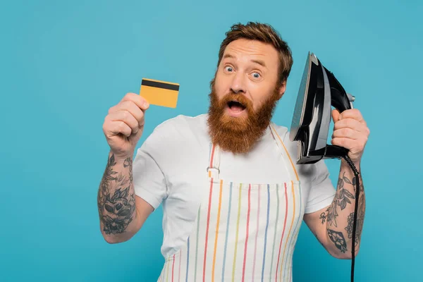 impressed bearded man with open mouth holding iron and credit card isolated on blue