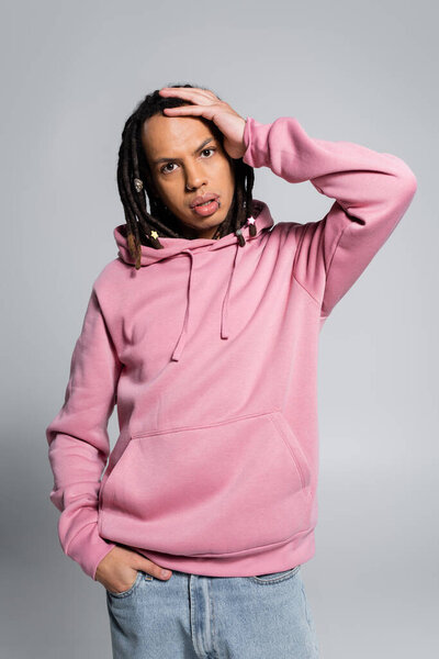 confused multiracial man with dreadlocks posing with hand in pocket on grey 