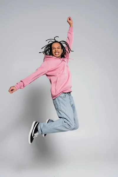 stock image full length of positive multiracial man with dreadlocks jumping with outstretched hands on grey 