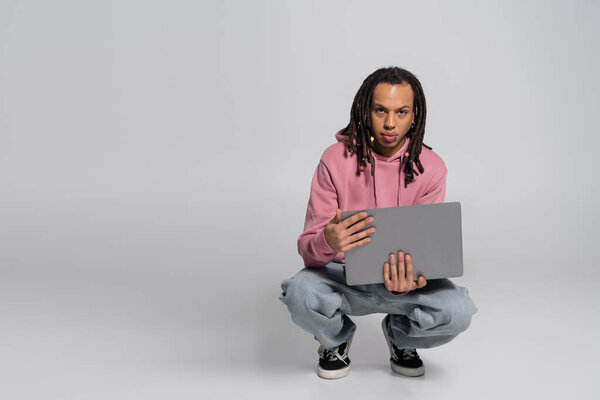 multiracial man in pink hoodie holding laptop while sitting on haunches on grey 