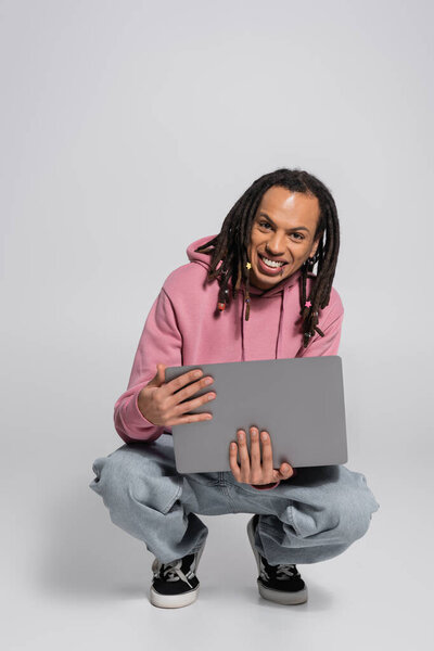 happy multiracial man in pink hoodie holding laptop while sitting on haunches on grey 