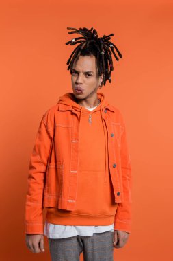 pierced multiracial man with dreadlocks pouting lips while standing in denim jacket on coral background  clipart