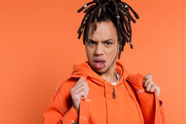 displeased multiracial man with dreadlocks adjusting denim jacket isolated on coral background 