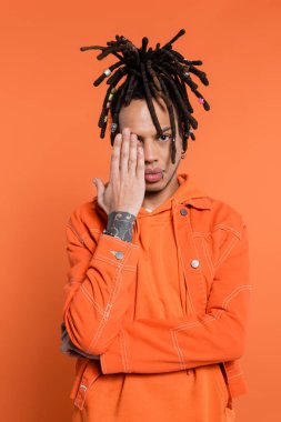 tattooed and multiracial man with dreadlocks covering half of face isolated on coral 