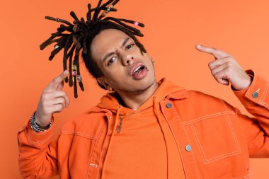 pierced and multiracial man with dreadlocks looking at camera while pointing with fingers isolated on coral background  clipart