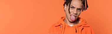 pierced and multiracial man with dreadlocks looking at camera while sticking out tongue isolated on coral background, banner  clipart