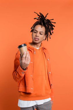 tattooed and multiracial man with dreadlocks looking at paper cup with coffee to go isolated on coral 