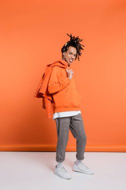 full length of positive multiracial man in hoodie holding denim jacket on coral background 