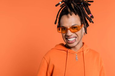 happy multiracial man with dreadlocks wearing stylish sunglasses isolated on coral background 