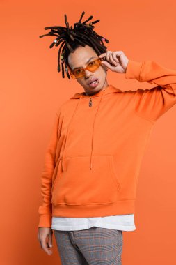 pierced multiracial man with dreadlocks adjusting trendy sunglasses on coral background 