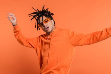 multiracial man in stylish sunglasses posing with opened mouth isolated on coral background 