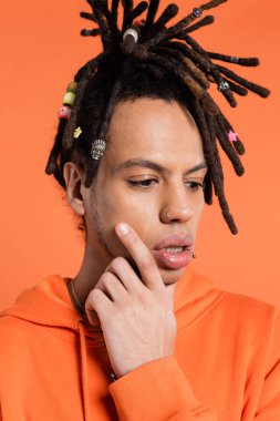 portrait of multiracial man with dreadlocks touching face while thinking isolated on coral background 