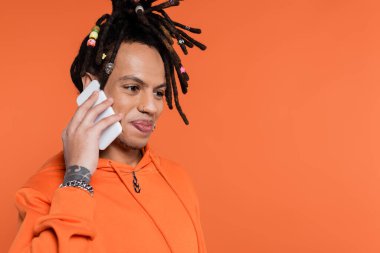 satisfied multiracial man with dreadlocks standing in hoodie and talking on smartphone isolated on coral 
