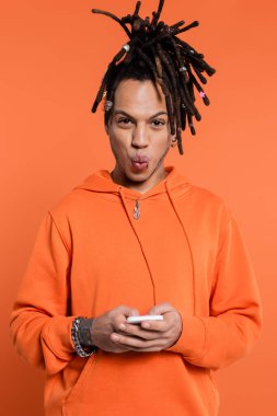 tattooed multiracial man with dreadlocks pouting lips and holding cellphone isolated on coral background 