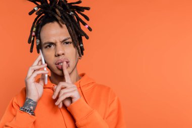 multiracial man with dreadlocks holding finger near lips and talking on cellphone isolated on coral  clipart